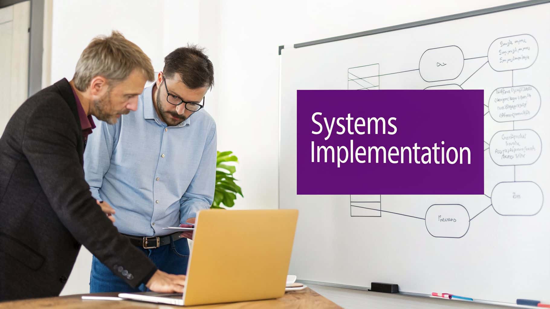 Two men collaborate on a laptop, discussing systems implementation with a whiteboard flowchart in the background.