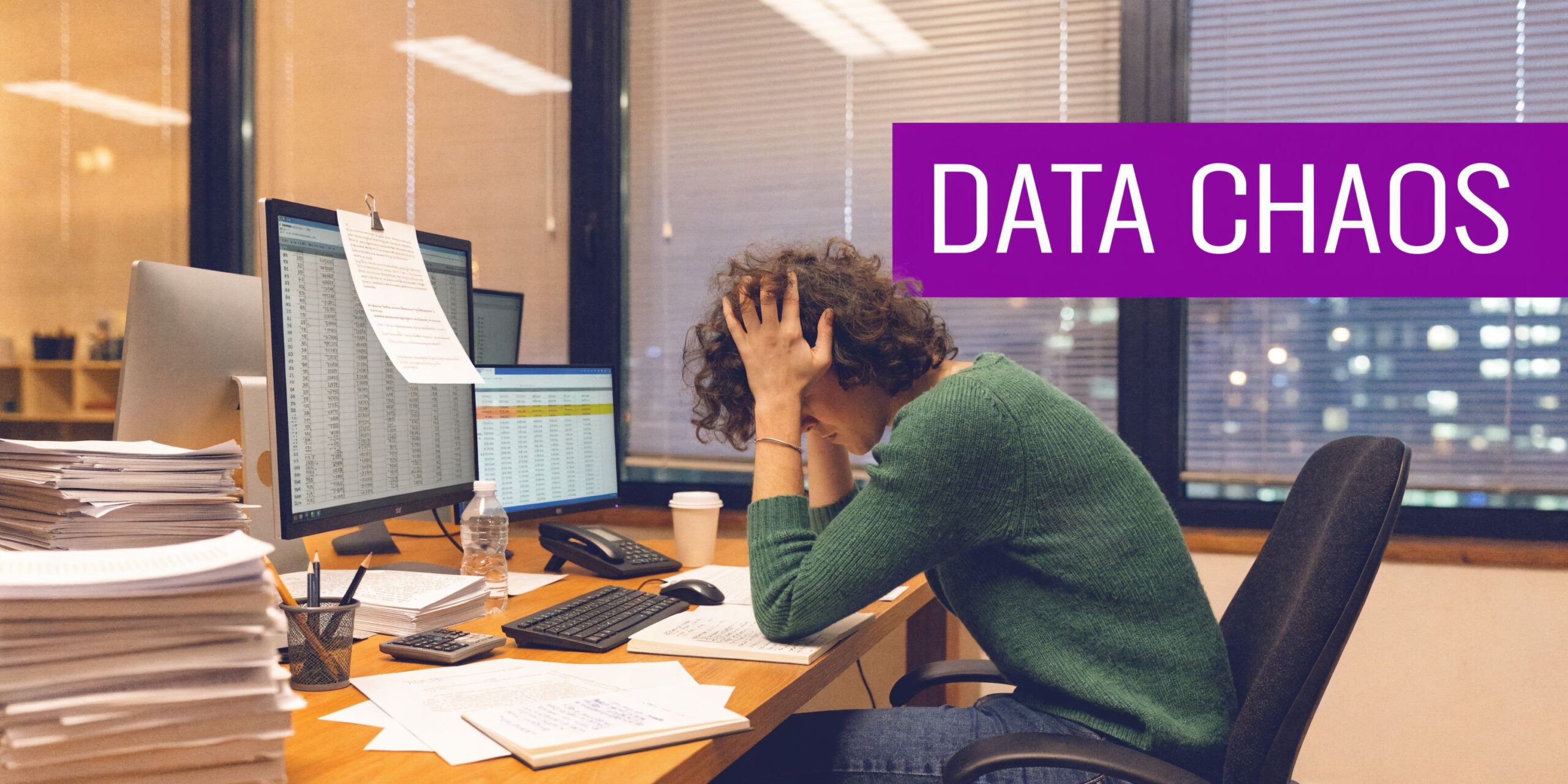A person sitting at an office desk feeling overwhelmed while looking at stacks of paperwork and computer screens.