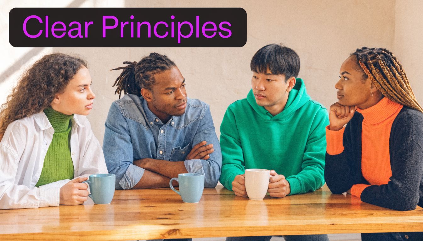A diverse group of four young adults sitting at a wooden table discussing ideas while holding mugs.