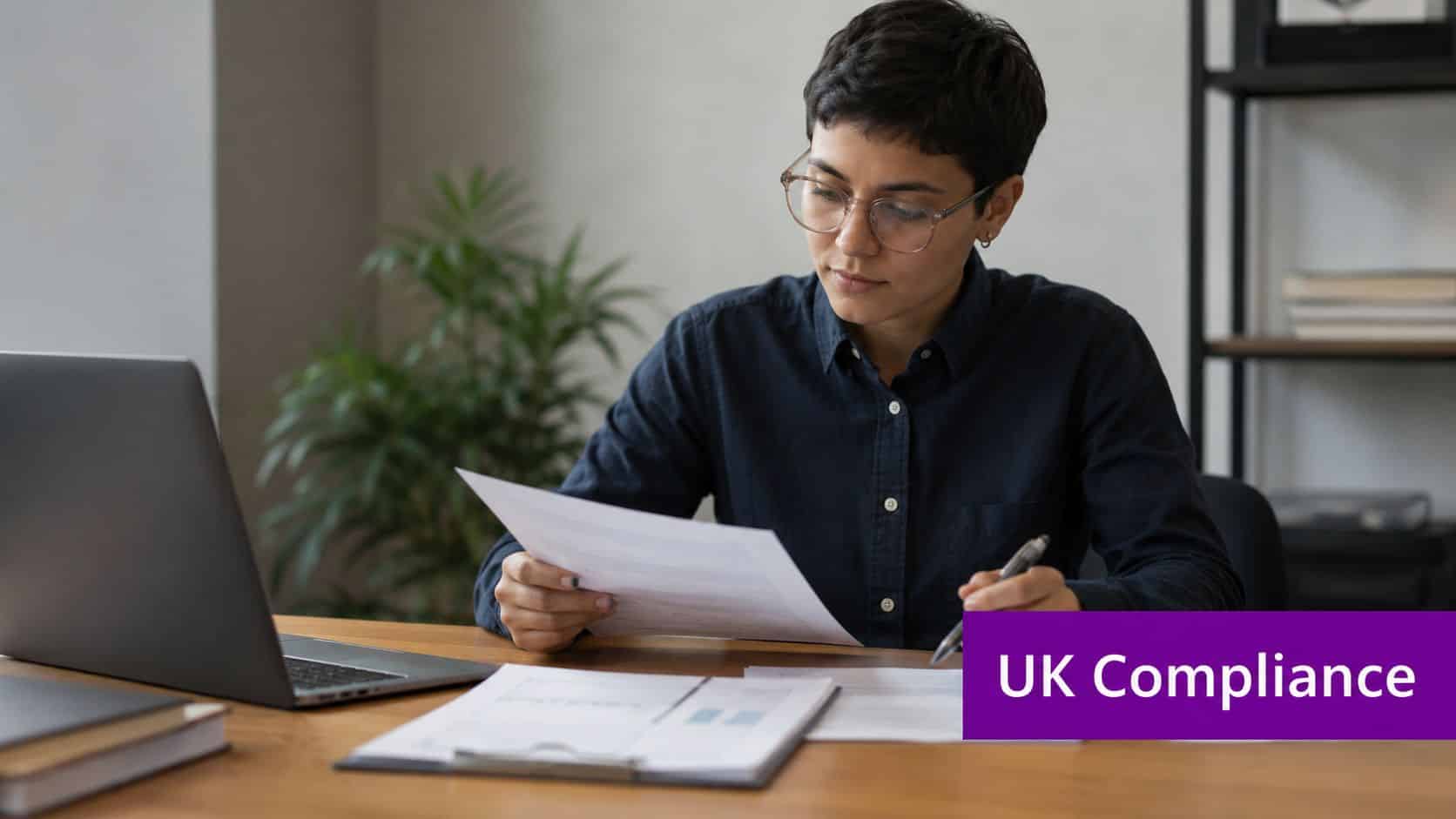 A professional woman wearing glasses reviewing documents at her desk with a laptop for business compliance.