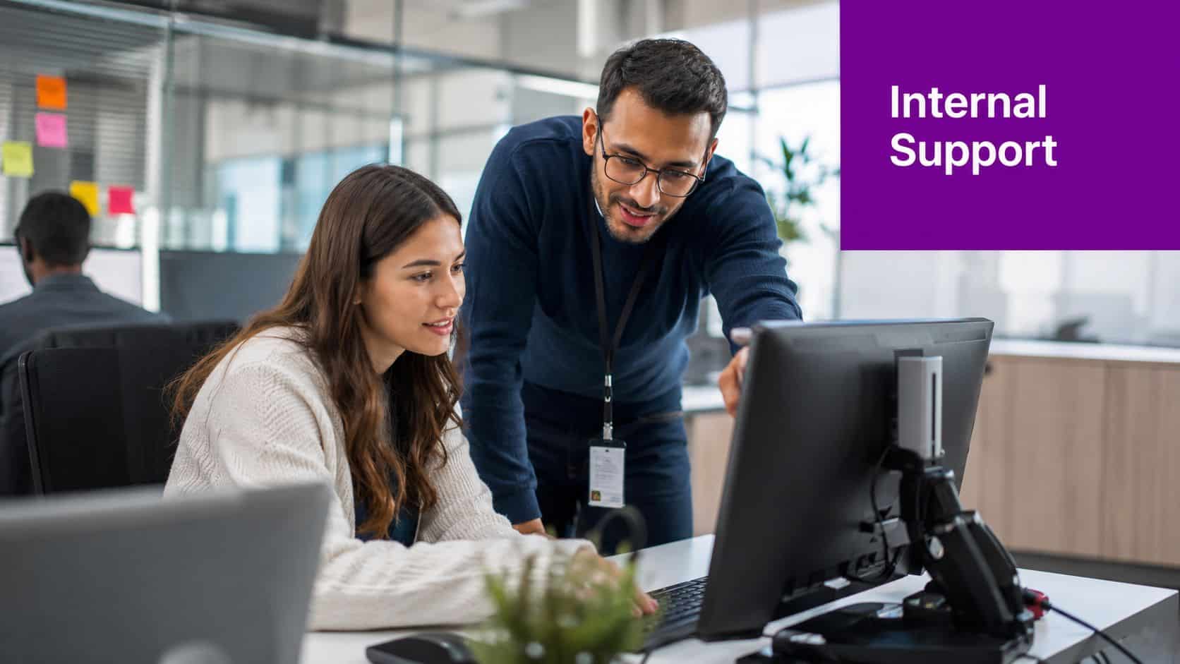 A male office colleague points at a computer monitor to assist a female coworker with work tasks.